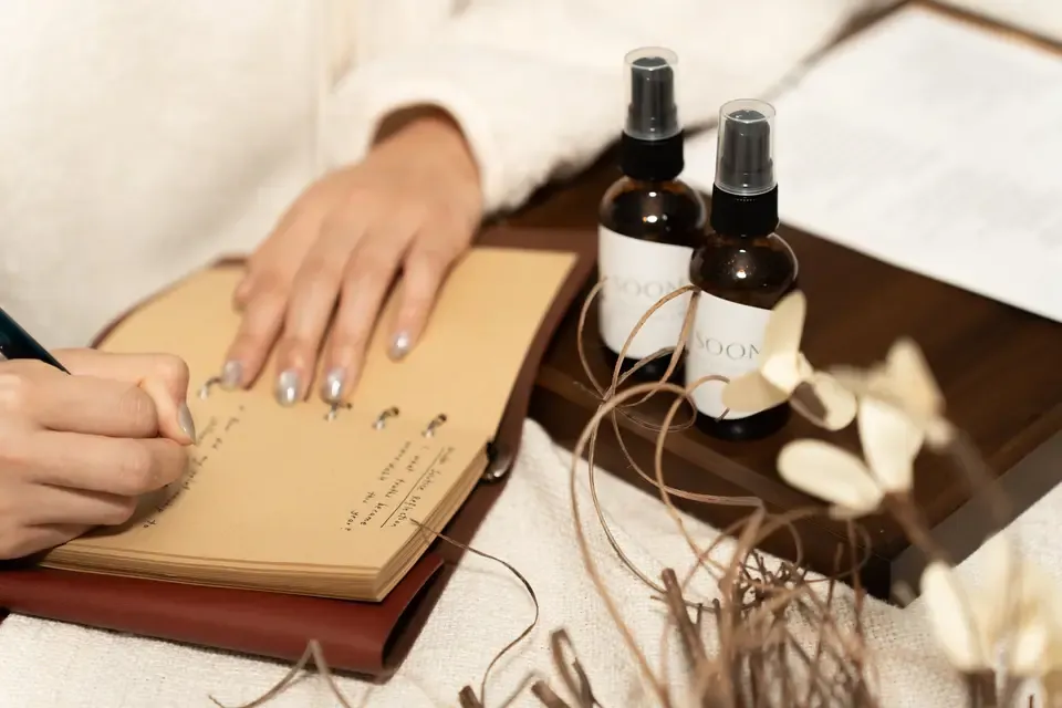 A person writing in a notebook with a pen, with two spray bottles labeled 'SOOM' and some decorative dried flowers nearby on a wooden tray.