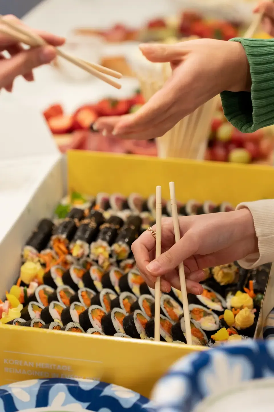 Close-up of hand holding chopsticks over a tray of sushi rolls at a food gather, with another person reaching for food in the background.