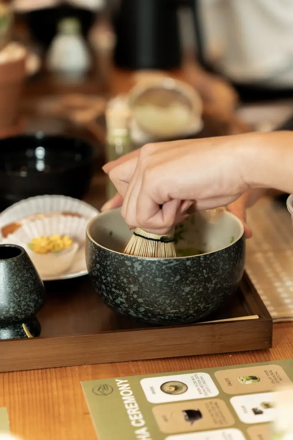 A person using a bamboo whisk to prepare matcha tea in a black speckled bowl on a tray, surrounded by small dishes and utensils on a wooden table.