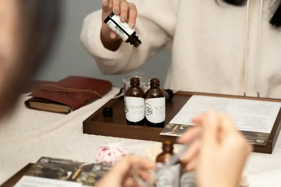 Person holding a small brown bottle above a tray with more brown bottles, with another person in the foreground taking a drop from a similar bottle, a notebook, and a clipboard on a table.