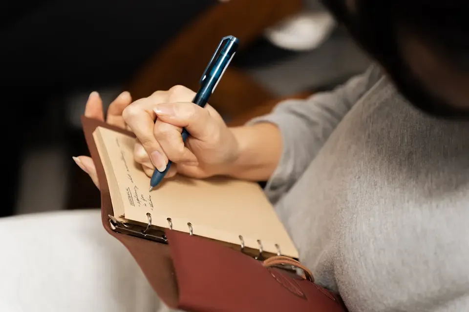 A person writing in a small notebook with a blue pen, holding the notebook open with one hand.