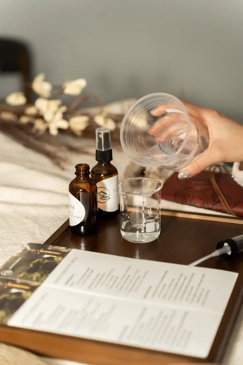 A person pours water from a clear plastic cup into a glass beaker on a wooden table. There are two brown bottles, a paper brochure or menu, and a white spray bottle nearby. A decorative branch with white flowers is in the background.