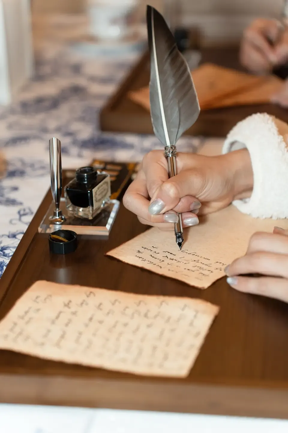 Person writing a letter with a quill on parchment paper, with ink bottle, on a wooden desk.
