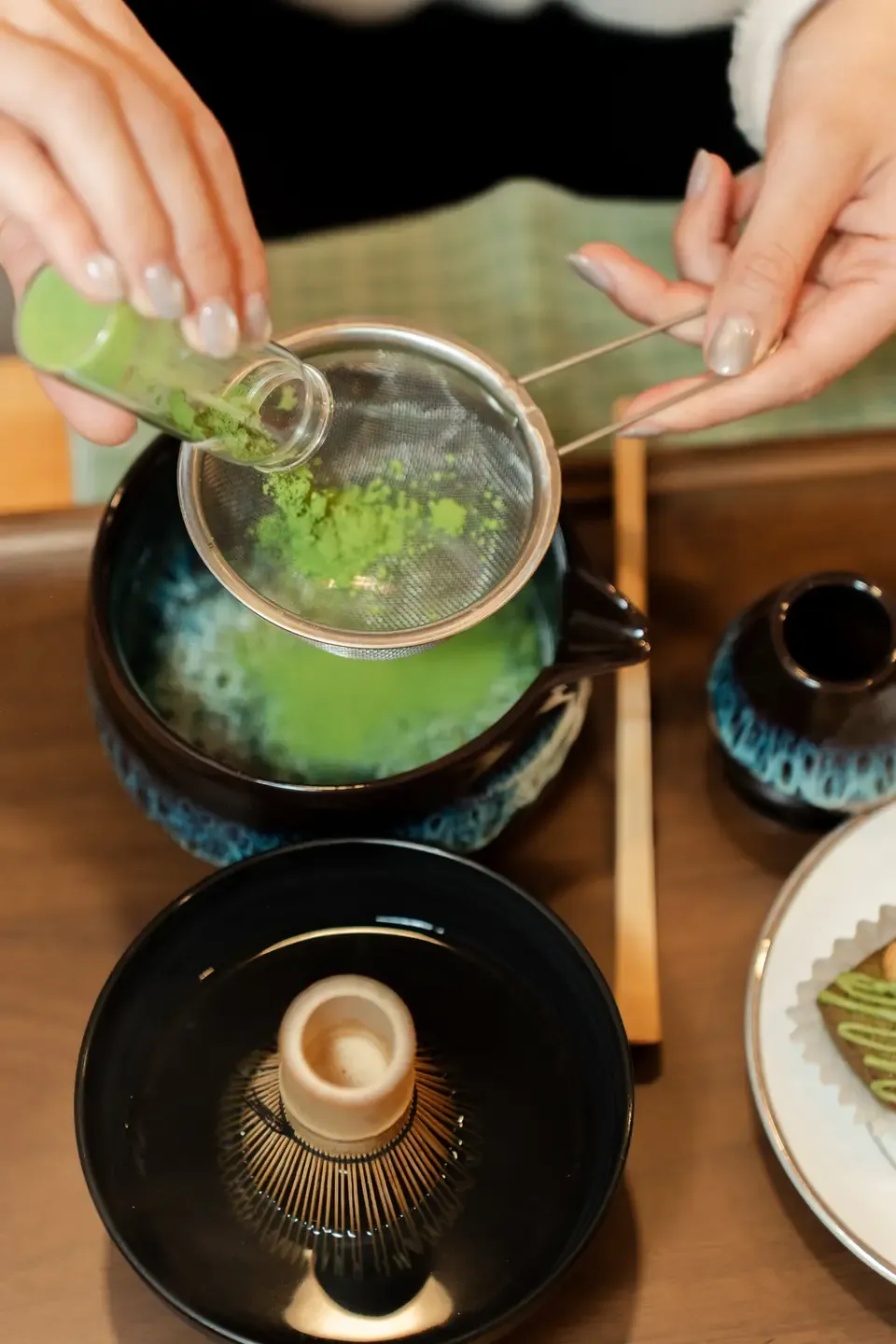 A person preparing matcha tea using a fine mesh strainer, pouring green powdered matcha through the sieve into a teapot. Various tea accessories and a plate with a green-topped dessert are also visible.