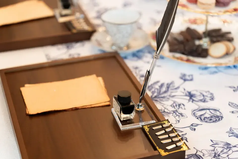 An inkwell with a quill pen on a wooden desk, with a folded piece of parchment paper and a set of fountain pen nibs nearby, on a floral-patterned tablecloth.