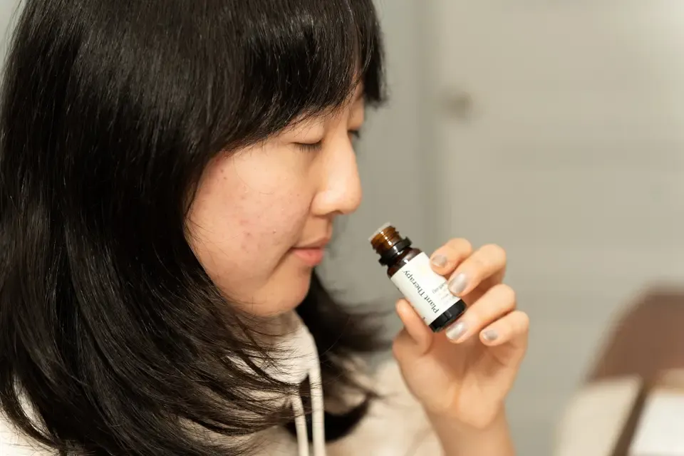 Young woman with dark hair and clear skin holding a small amber bottle labeled 'Plant Therapy' near her nose, appearing to smell it.
