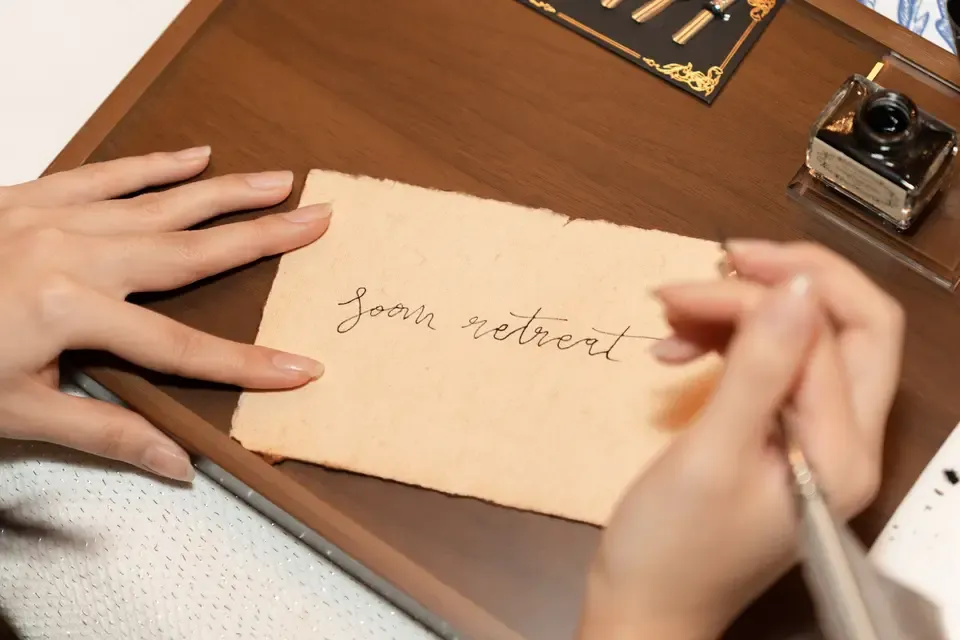 Person writing 'Soom retreat' on a piece of aged, torn paper with a pen, sitting at a wooden desk with ink bottle and calligraphy tools.