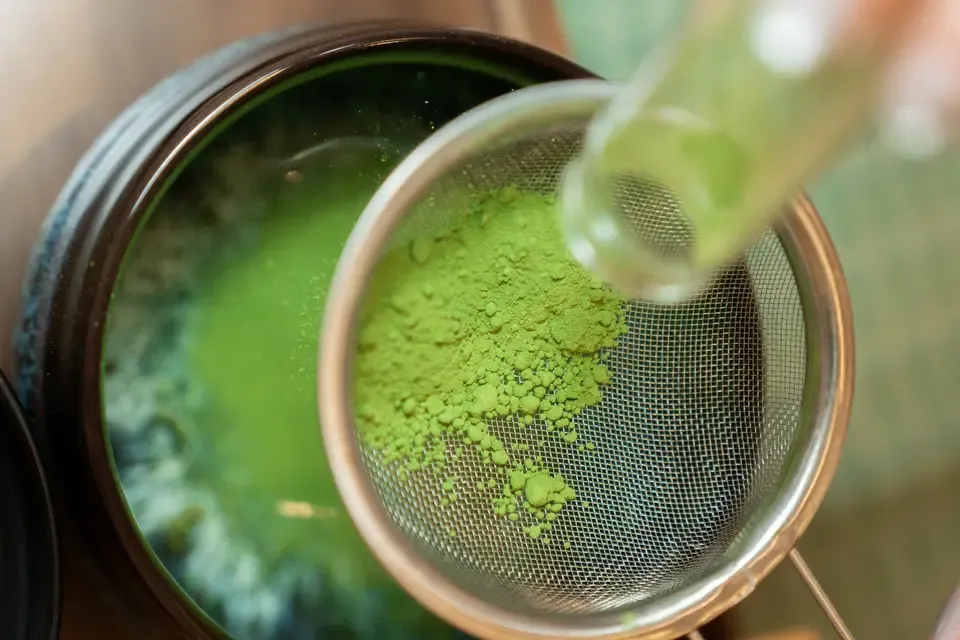 Green matcha tea powder being sifted through a metal strainer.