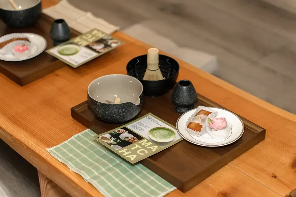 Table setting with a tray holding a black bowl with a bamboo container, a white cup, and a small black vase, along with a plate containing two desserts, in a cozy restaurant or cafe.