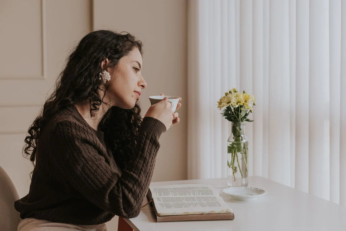 Someone with a cup of tea and an open book at a quiet table beside a bright window.