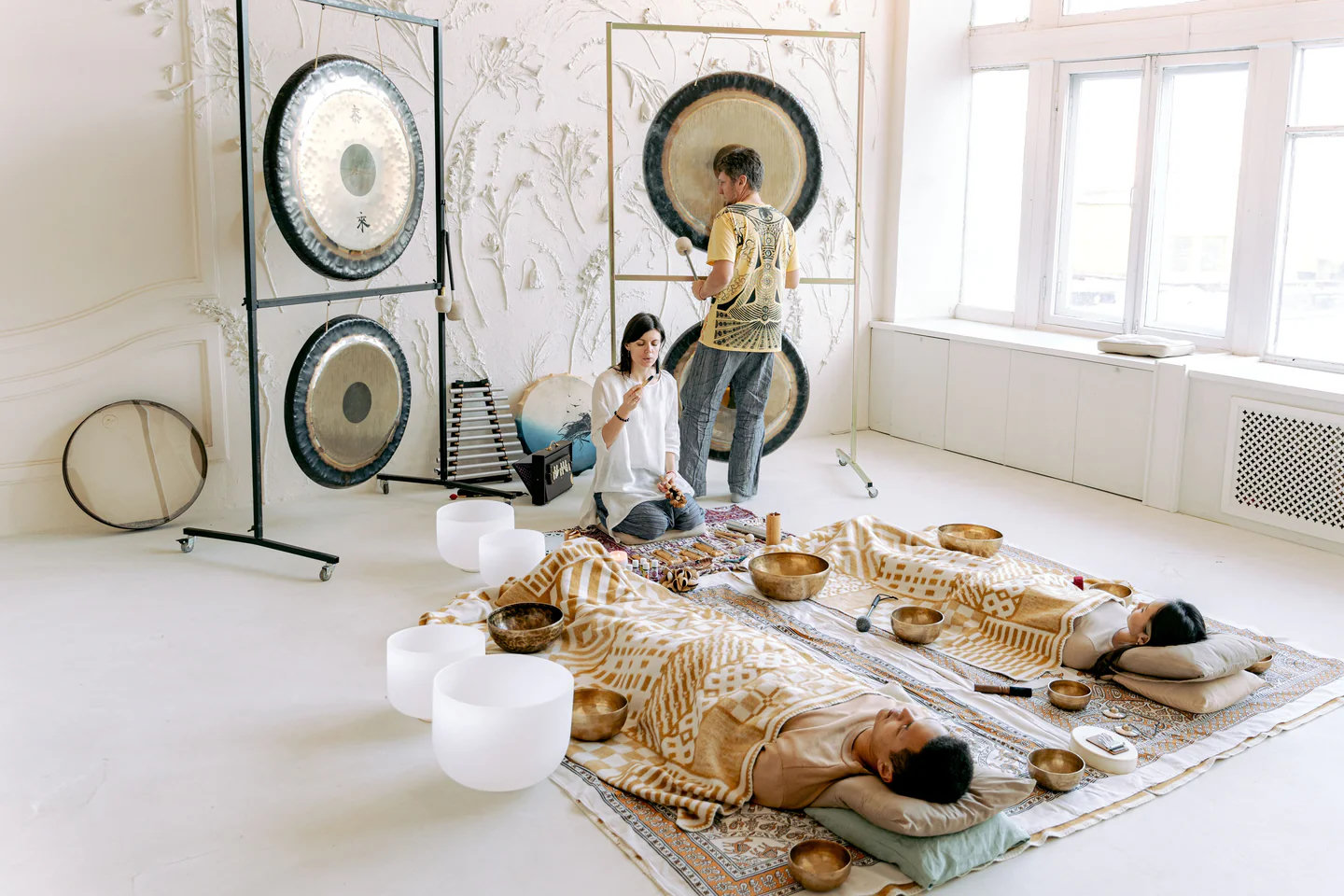 People resting under blankets on floor mats among singing bowls and gongs in a bright room.