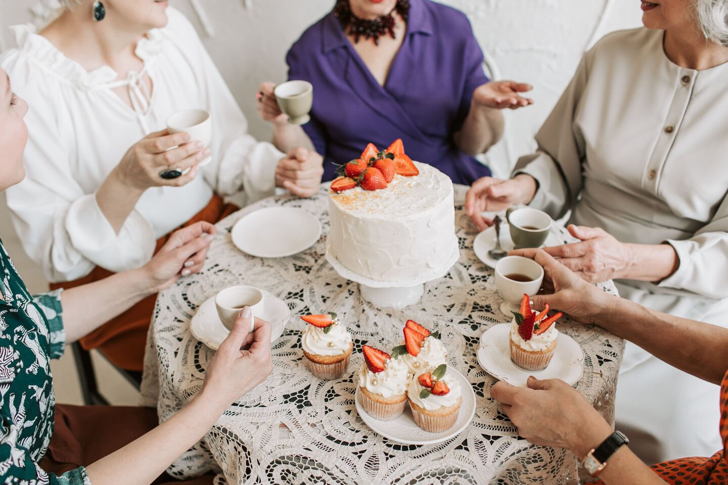 A small group around a lace-covered table in soft daylight, sharing tea, a strawberry-topped cake and cupcakes.